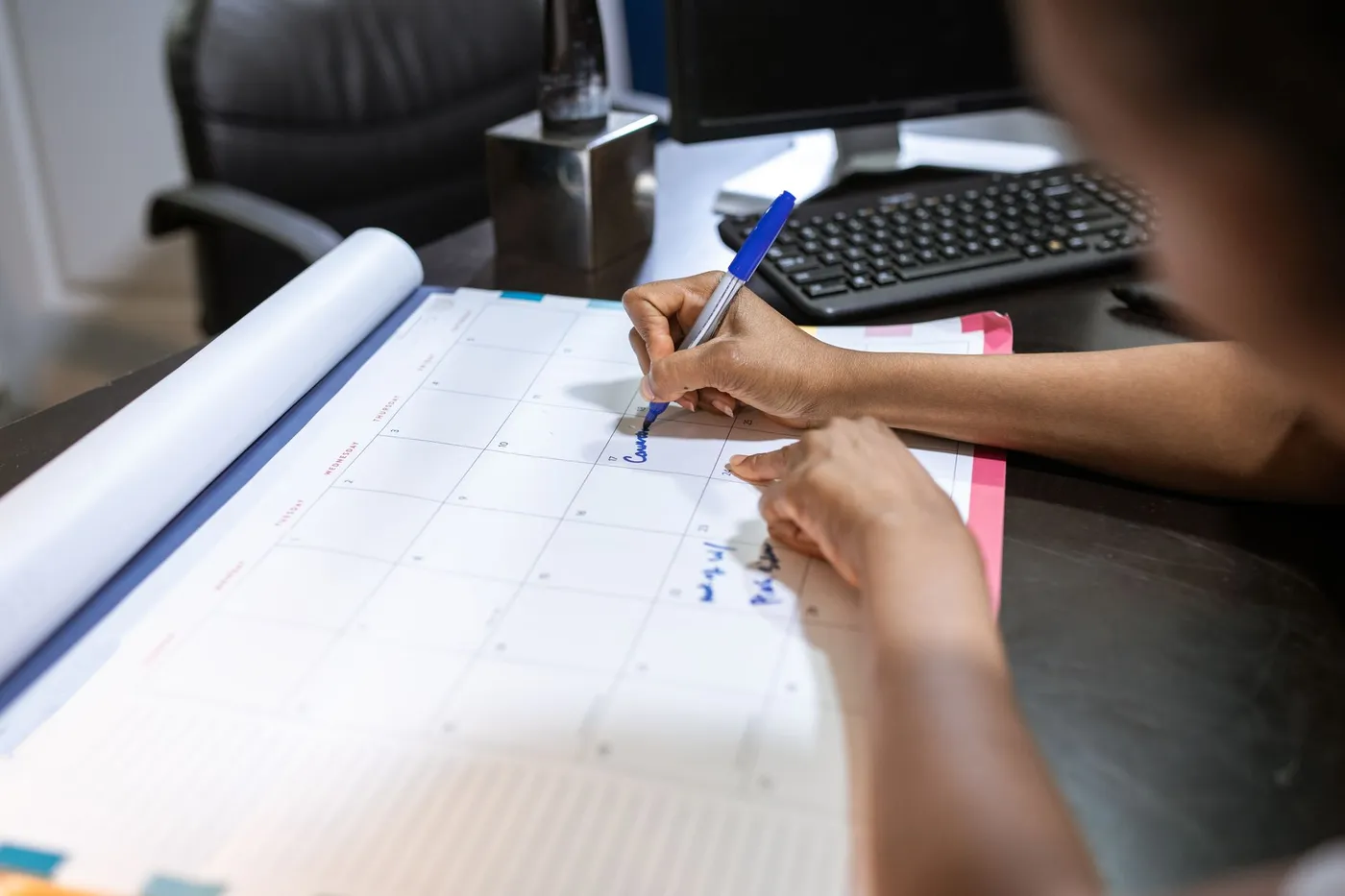 A person writes in blue marker on a large monthly desk calendar next to a computer keyboard, planning dates weeks ahead.