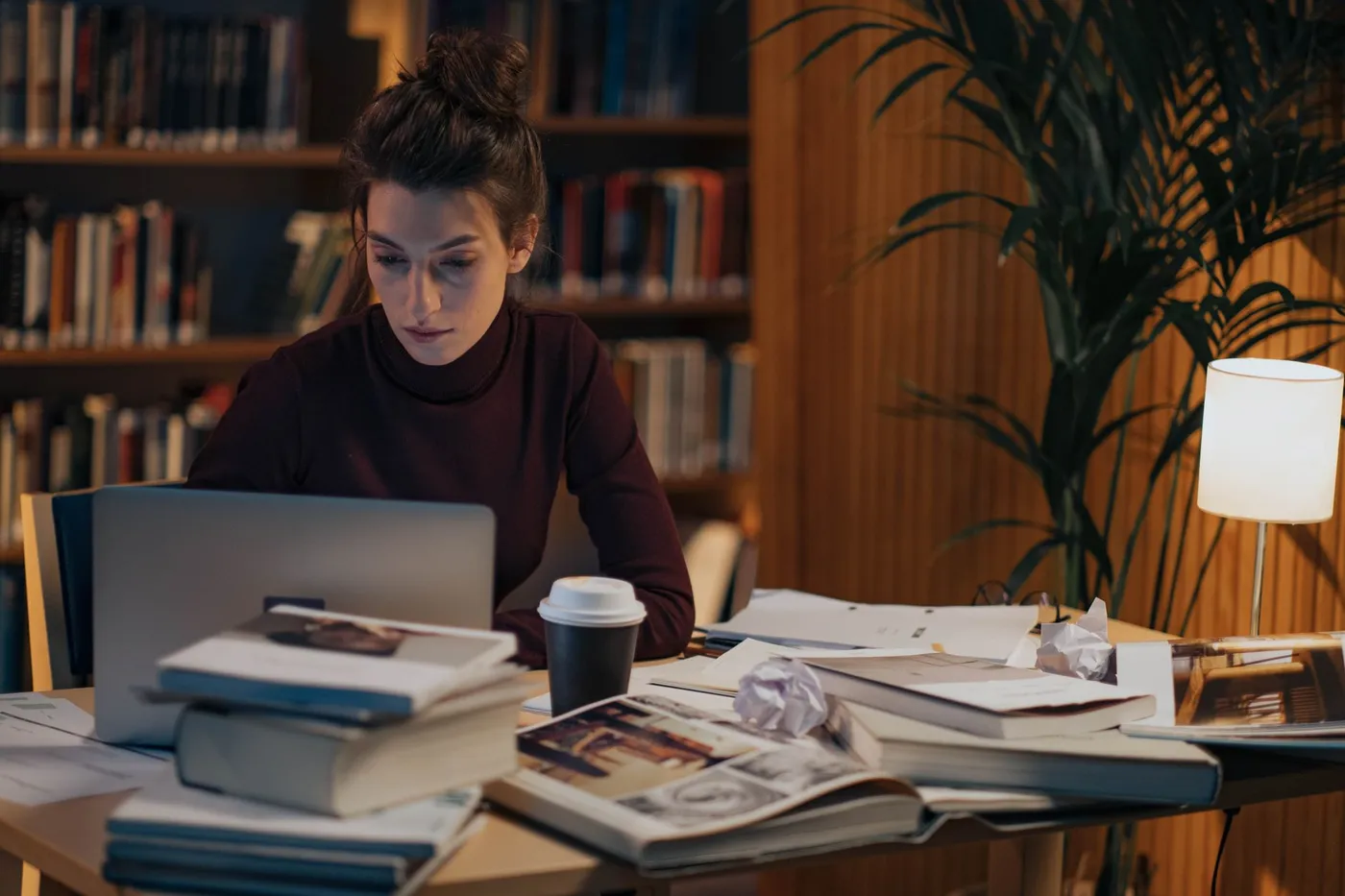 A woman in a dark red turtleneck works at a laptop late in the evening, a takeaway coffee cup and stacks of books and loose papers spread across the desk beside her, with filled bookshelves behind.