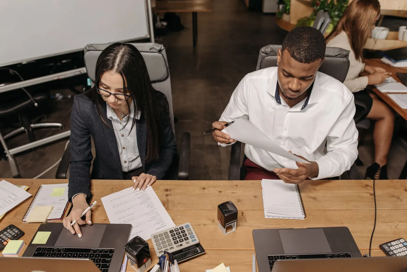 Seen from above, two young professionals work side by side at a wooden desk: a woman in glasses writes beside her laptop and a printed balance sheet, while a man in a white shirt reads a page he holds up, with calculators and a desk stamp between them.