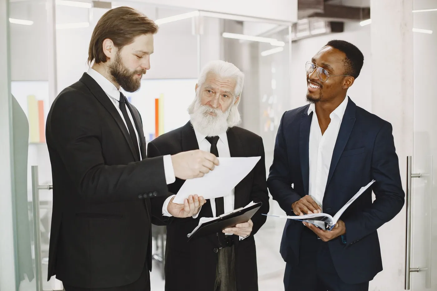 Three business professionals in suits reviewing printed documents together at an office table